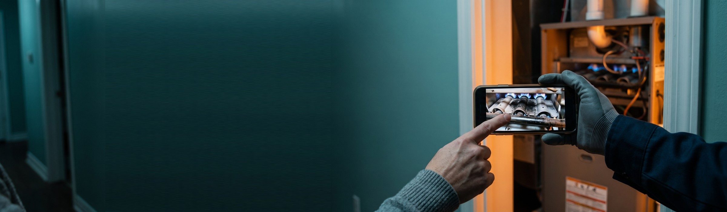 A technician's gloved hand holds out a phone showing a photo of sooty furnace burners while a homeowner points at the screen, in front of an open furnace closet
