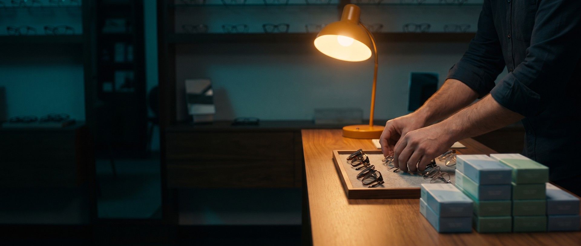 After hours in a dark optical shop, an optician's hands set eyeglass frames on a display tray under a single amber desk lamp beside stacks of fresh contact lens boxes
