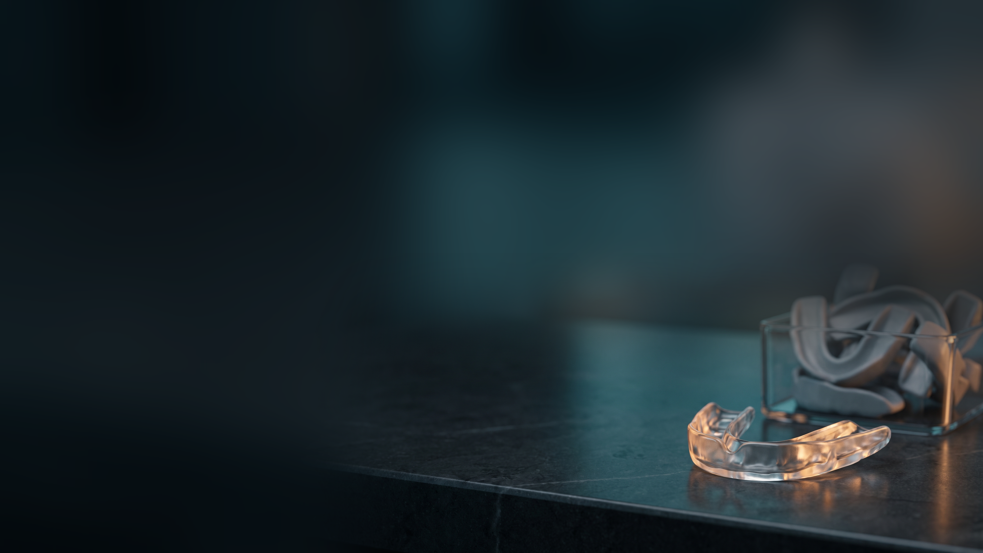 A clear custom-molded dental night guard glowing on a dark counter beside a bin of identical generic mouthguards