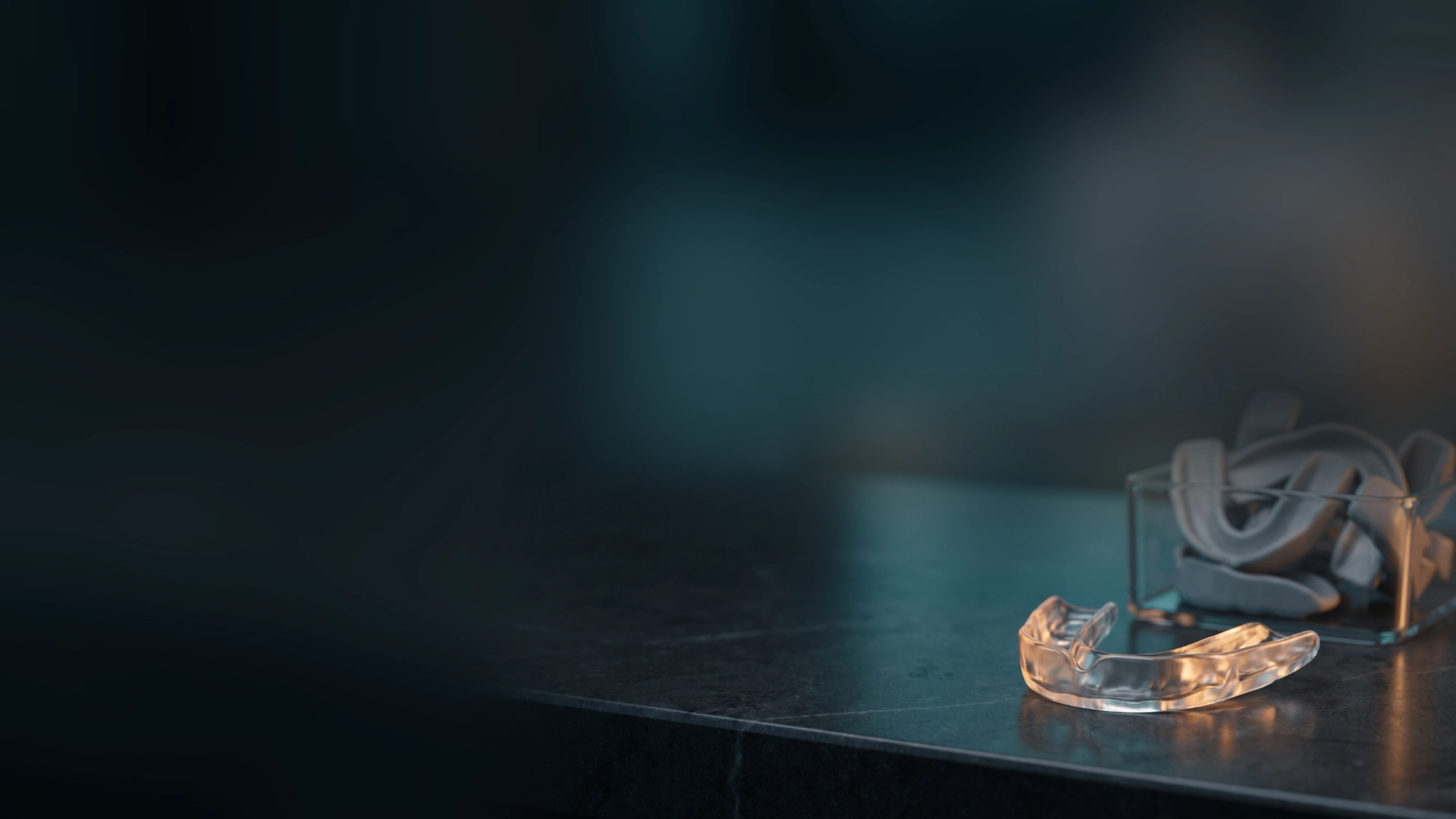 A clear custom-molded dental night guard glowing on a dark counter beside a bin of identical generic mouthguards