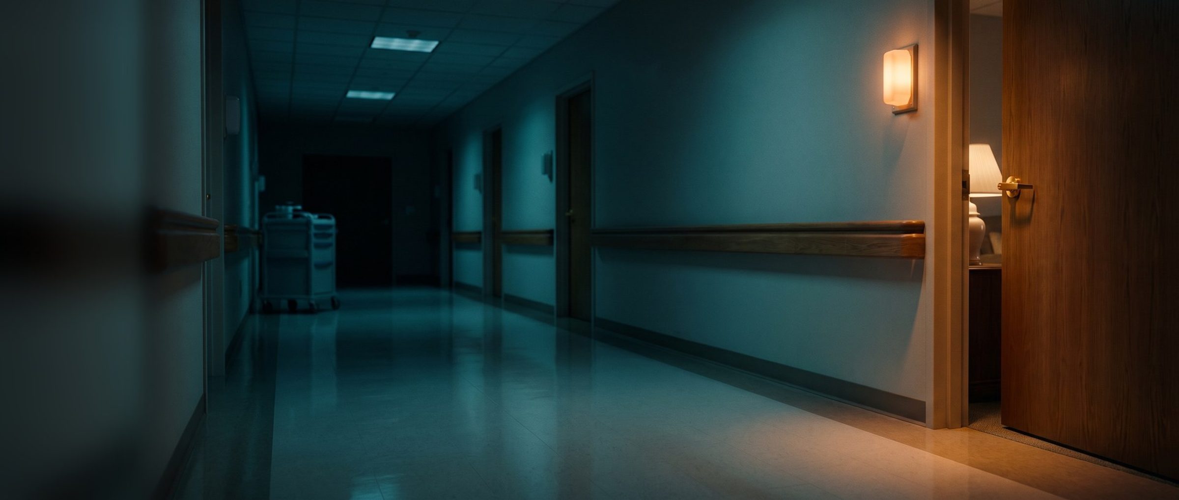 A nursing home corridor at night in cool shade, one resident's door standing ajar with a lamp on inside and the call light beside it lit amber while the others down the hall stay dark, a medication cart parked far down the empty hall
