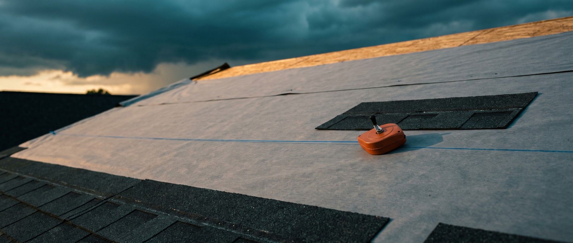 A roof deck mid-reroof in late light: an orange chalk reel rests on gray underlayment beside a crisp blue chalk line snapped across the deck, new charcoal shingles along the eave and dark storm clouds building behind the ridge