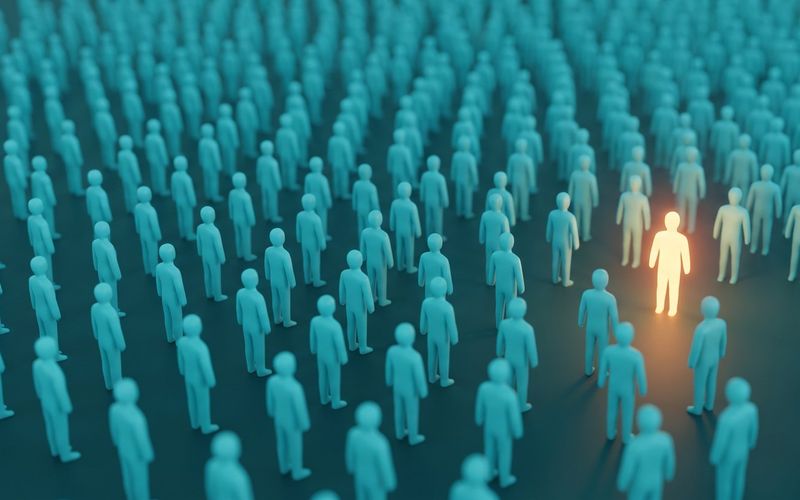 A vast crowd of identical teal figures on a dark floor, with one single figure lit warm amber standing out among them