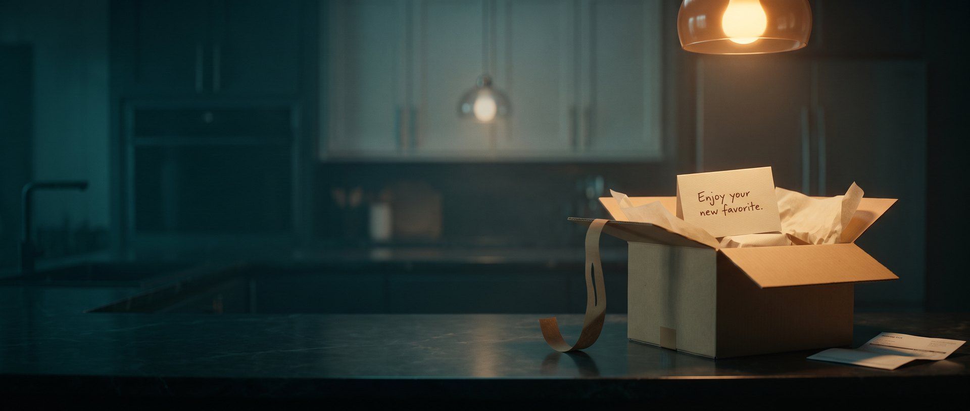 An opened kraft shipping box on a dark kitchen counter, tissue paper pulled back and a handwritten card inside reading "Enjoy your new favorite," with a packing slip beside it under a warm pendant light