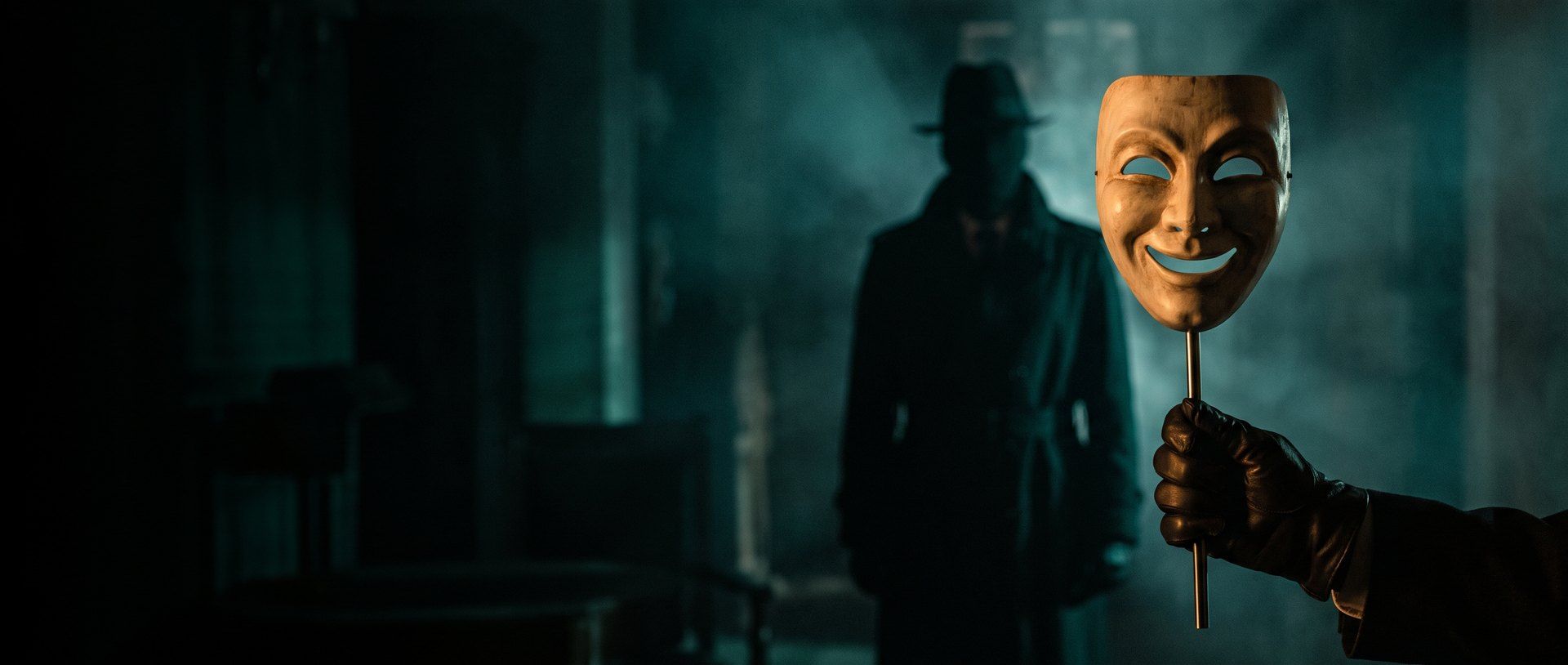 A gloved hand holding up a smiling theater mask on a rod while a figure in a hat and long coat waits in the shadows behind it, lit in dark teal with a warm amber glow on the mask.