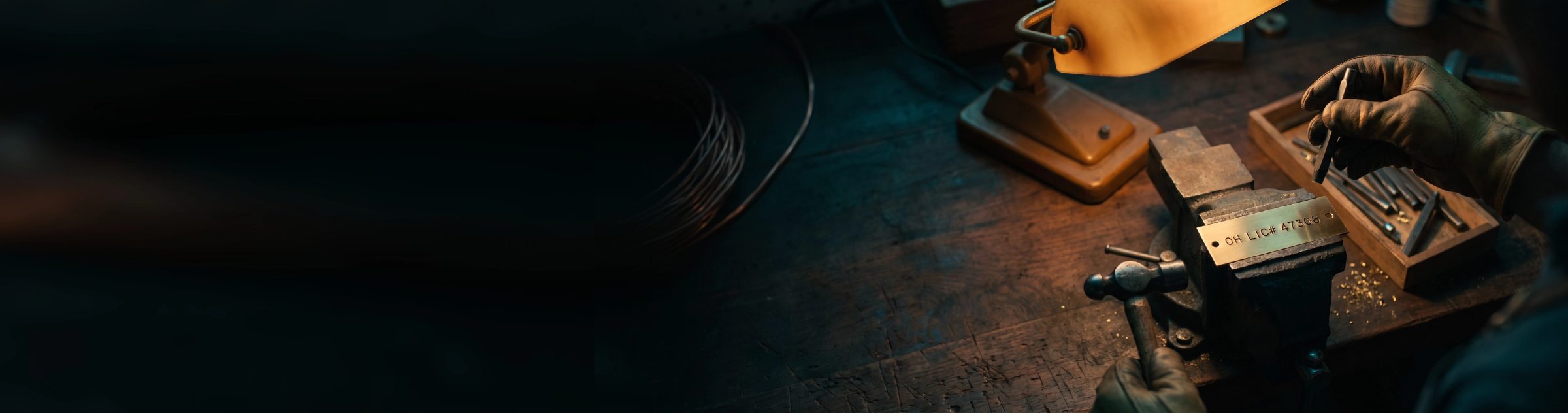 A gloved hand lifting a steel punch from a small brass plate held in a bench vise, the plate freshly stamped OH LIC# 47306, under a warm work lamp on a scarred workbench