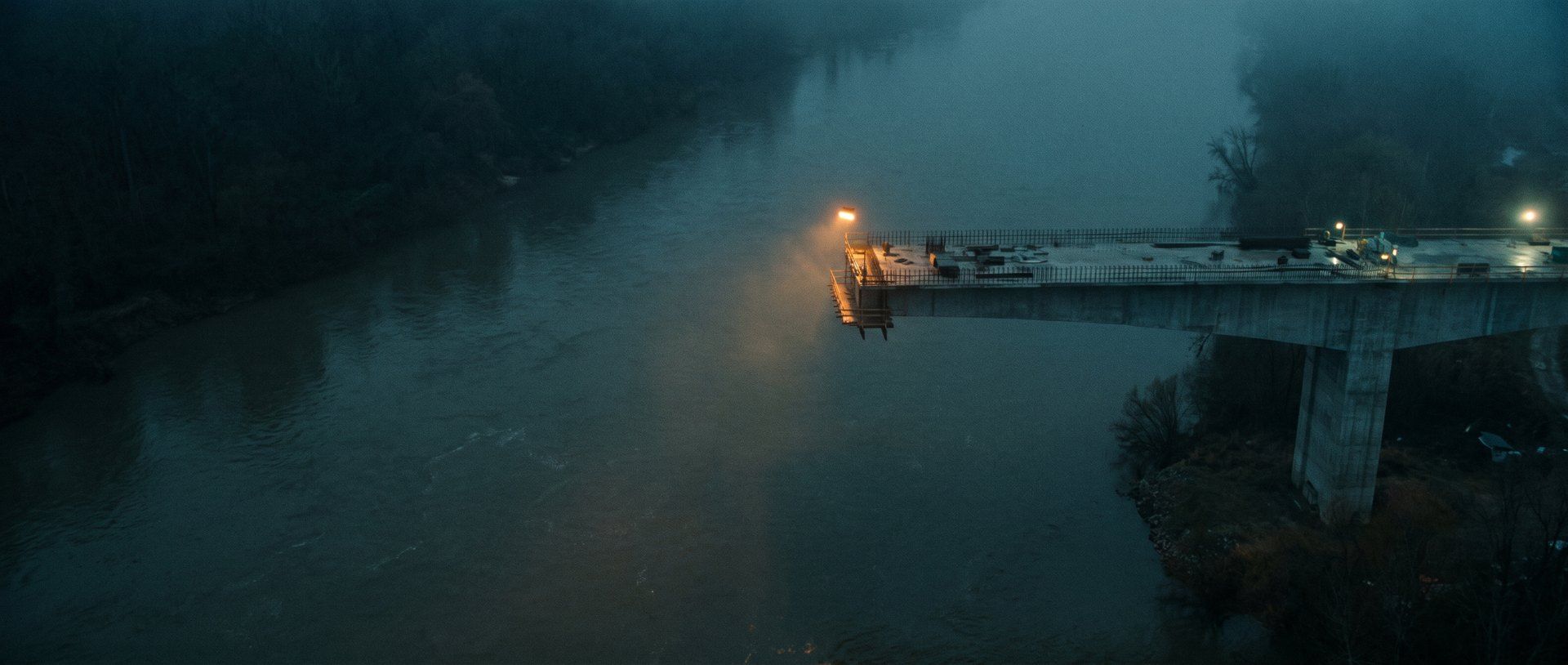 An unfinished road bridge stopping in mid-air over a river at dusk, with a single amber lamp at the cut end
