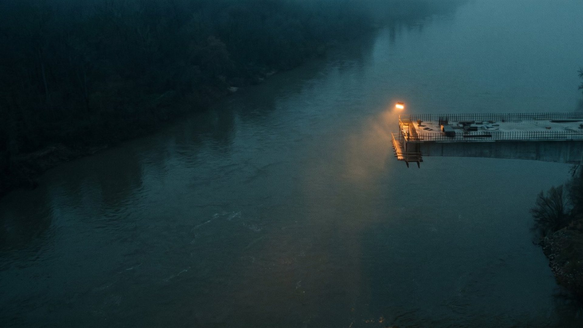 An unfinished road bridge stopping in mid-air over a river at dusk, with a single amber lamp at the cut end