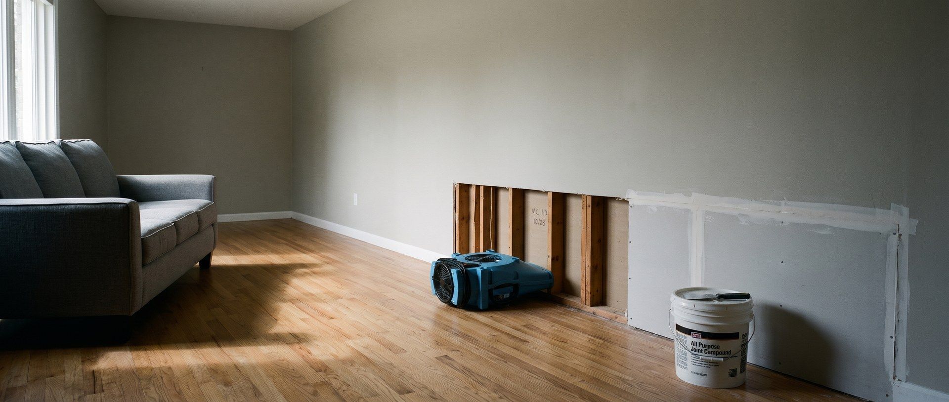 In a living room, the bottom of one wall is cut away to the studs with a blue air mover blowing into the opening, while the rest of the cut is already patched with new taped drywall and a bucket of joint compound sits below