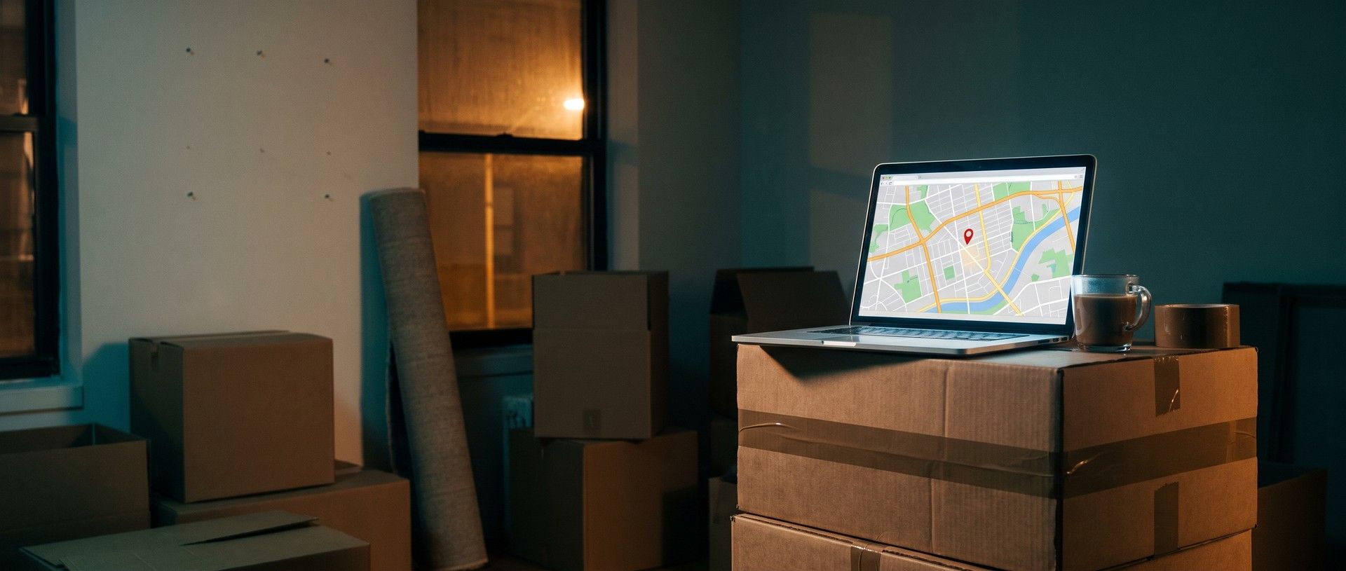 A half-packed apartment at night: taped moving boxes, a rolled-up rug and a bare wall with nail holes, and on a stack of boxes an open laptop glowing with a city street map and one red pin