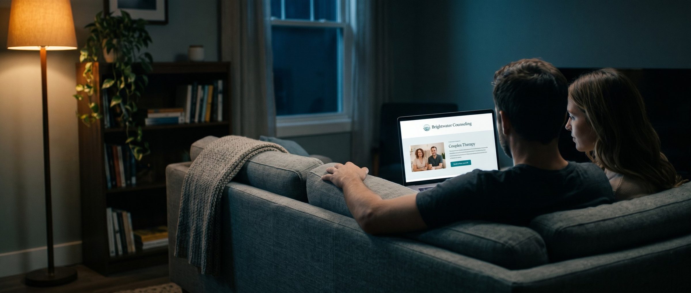 At night in a lamp-lit living room, a couple on a gray couch, seen from behind, read a counseling practice's Couples Therapy page on a laptop, with a photo of two therapists and a Book a free consult button