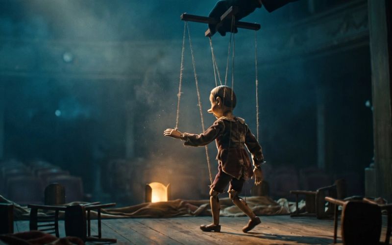 A wooden marionette performing on a dim stage, its strings running up to an unseen hand above