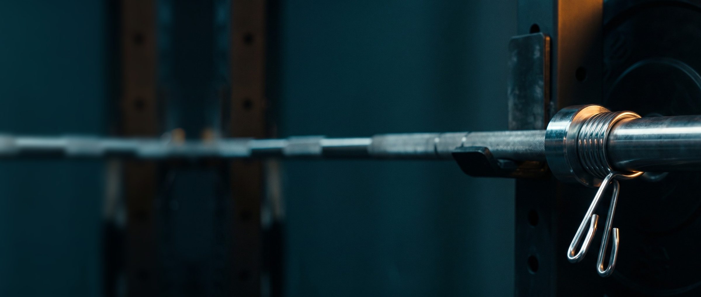 Close-up of a chrome spring collar clamped on a loaded barbell against black weight plates, its coiled spring and squeeze handles lit warm, the bar running off into a dark teal gym