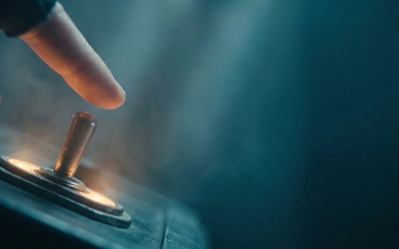 A fingertip about to flip a glowing brass toggle switch on a dark control panel, warm amber light around the switch, deep teal haze behind