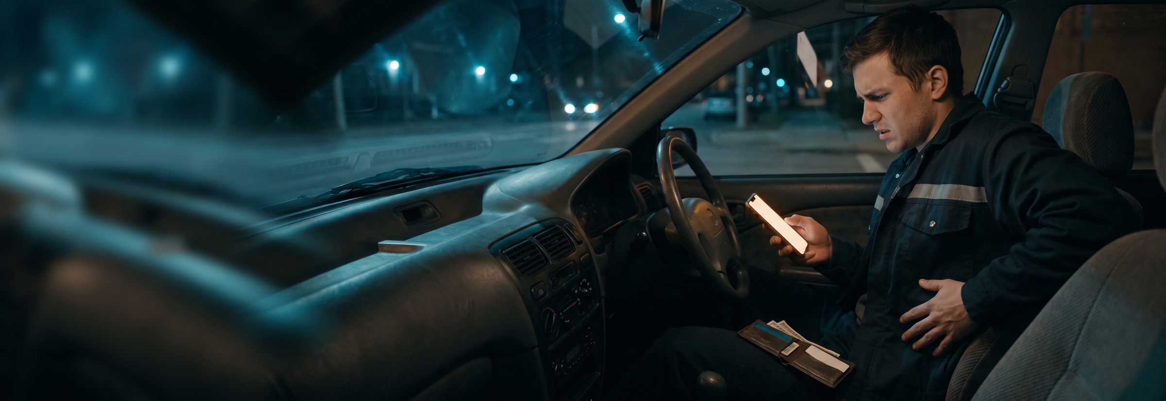 At night in a worn older car, a young worker in a reflective work jacket holds his side and stares at his phone, a thin wallet with a few bills in his lap, weighing what a visit will cost