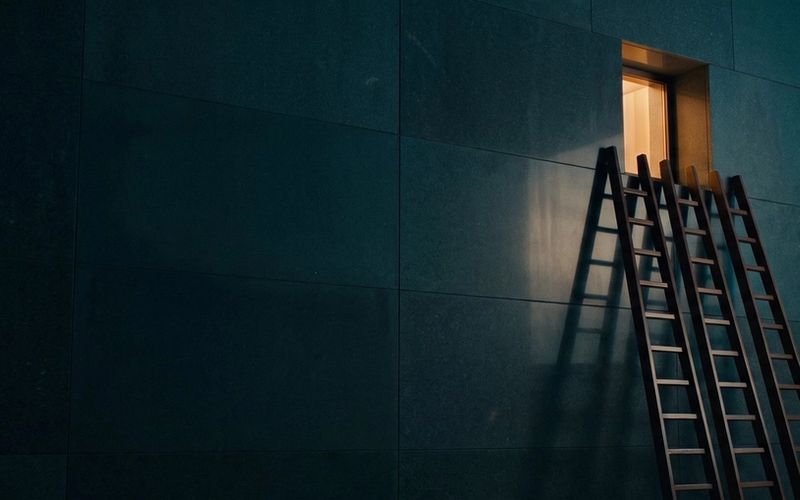 Three ladders leaning against the same single lit window in a dark wall