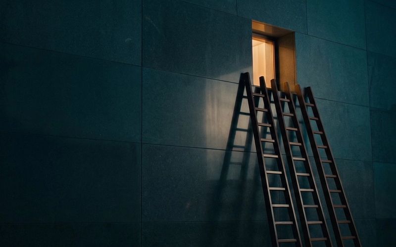 Three ladders leaning against the same single lit window in a dark wall