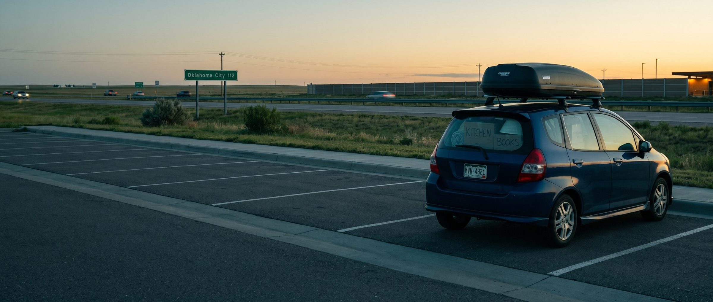 A small blue hatchback packed to the roof with moving boxes marked Kitchen and Books, a cargo box on top, parked at an interstate rest area at dawn, with a green highway sign in the distance reading Oklahoma City 112