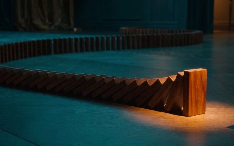 A long chain of dominoes toppled left to right but halted one tile short of the end, the final tile still standing under warm amber light - a checkout that runs almost to completion, then stops.