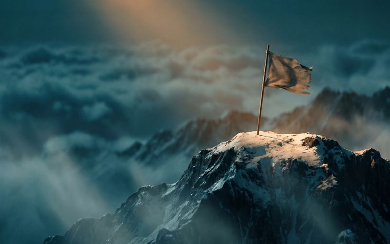 A single flag planted on a dramatic mountain summit at amber dawn, deep teal clouds in the valley below - the objective reached.