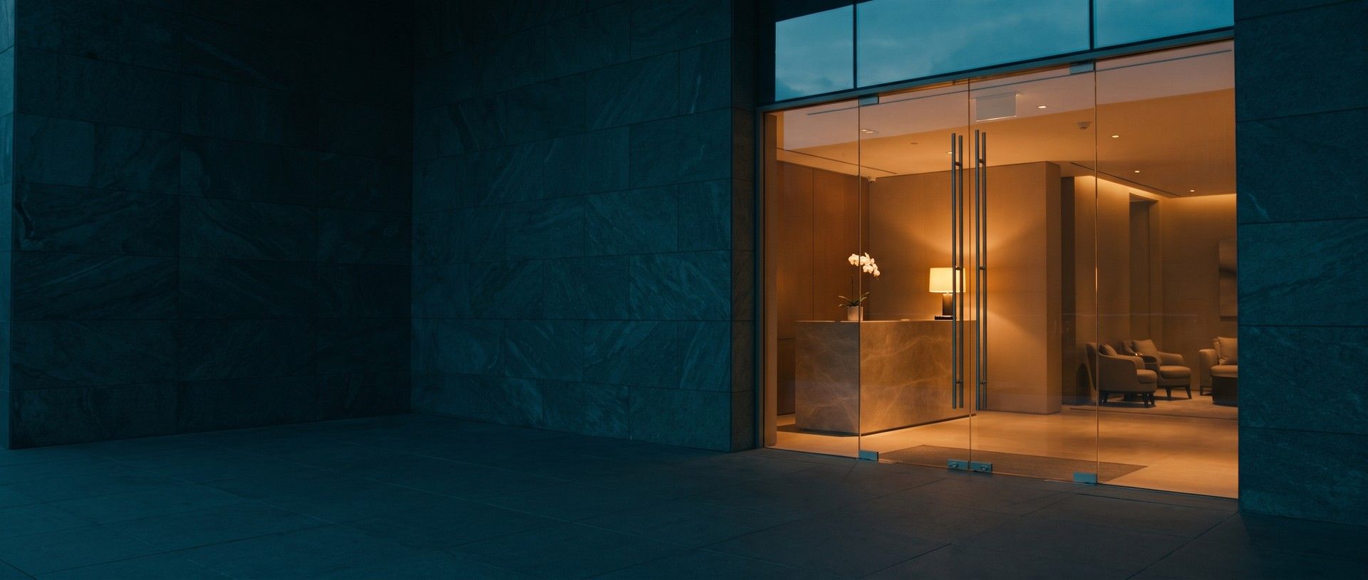 Glass entrance doors of a med spa at dusk, a stone front desk with a white orchid and a warm lamp glowing inside past a dark stone wall