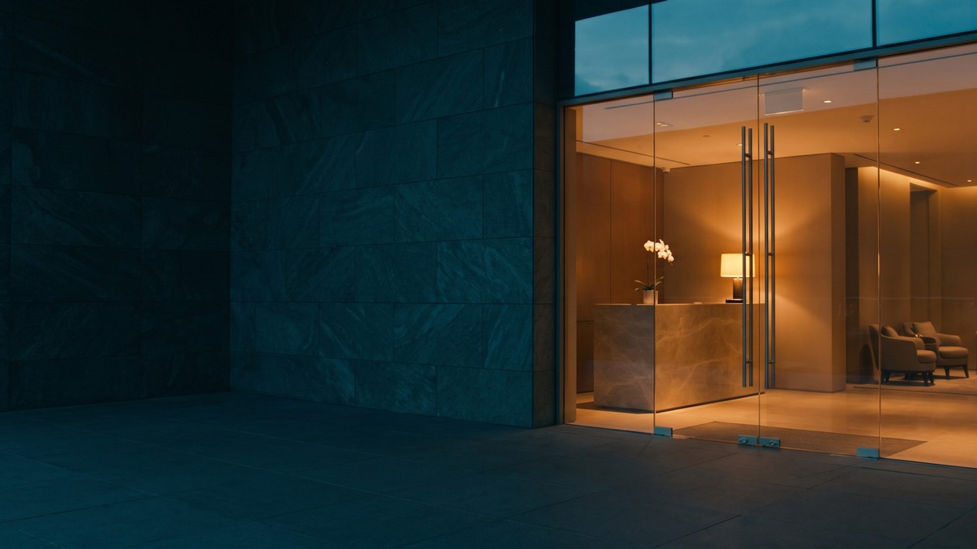 Glass entrance doors of a med spa at dusk, a stone front desk with a white orchid and a warm lamp glowing inside past a dark stone wall