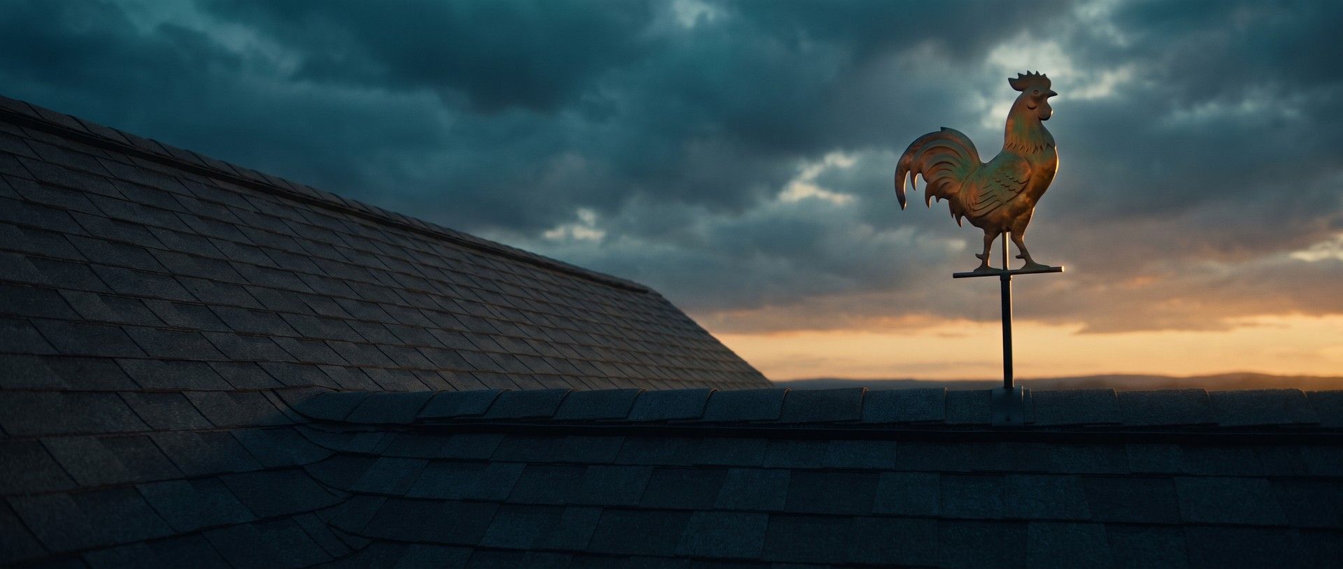 A copper rooster weathervane on a plain black rod stands on the ridge of a charcoal shingle roof, dark teal storm clouds breaking over an amber sunset behind it