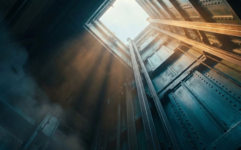 Looking straight up a deep industrial lift shaft that opens into bare sky at the top, with nothing capping it