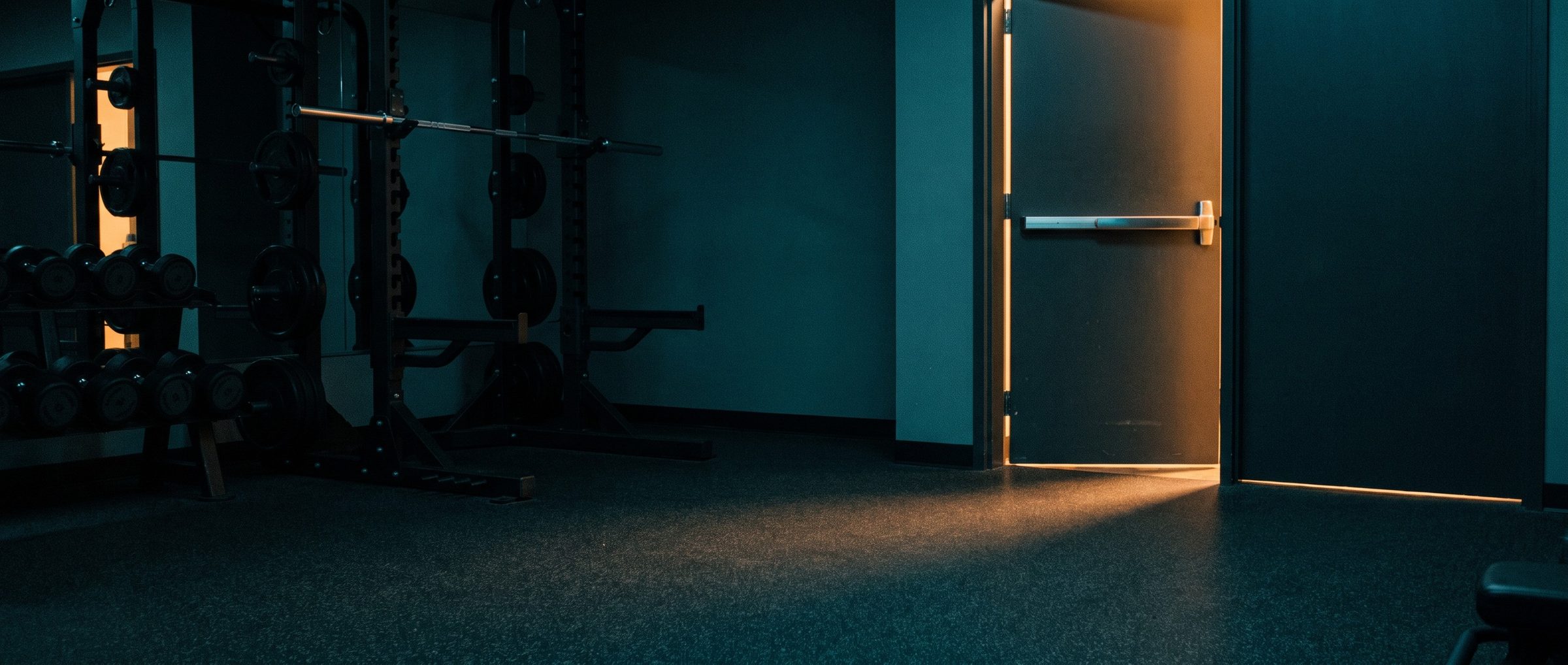 A steel gym side door with a chrome push bar standing slightly open at dusk, a line of warm evening light spilling across the dark rubber floor, squat racks and dumbbells in shadow