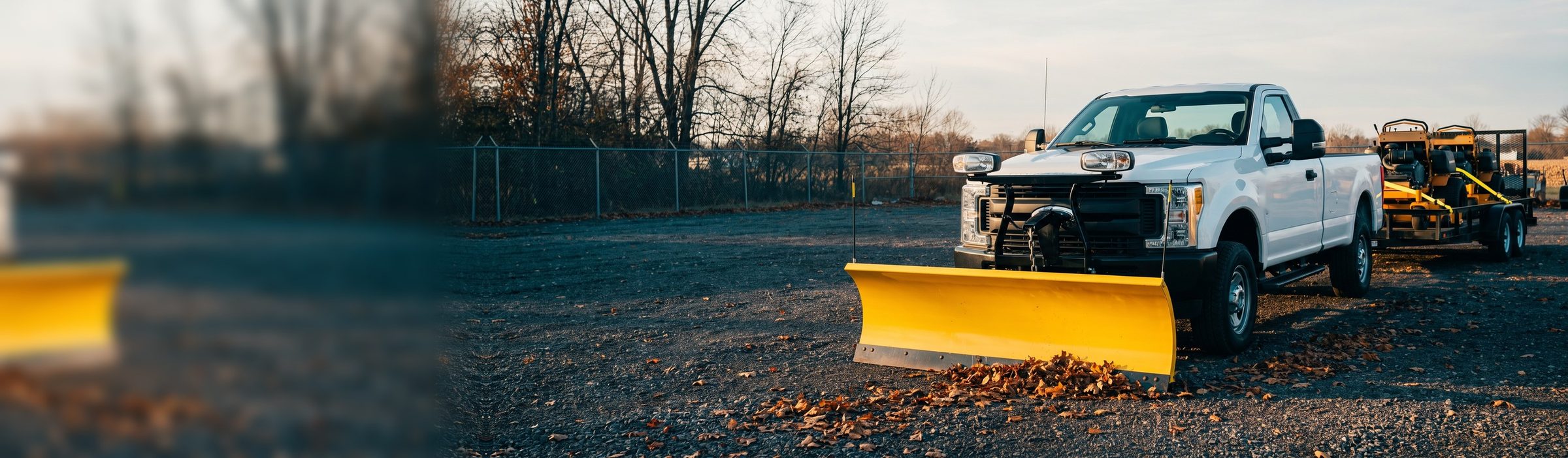 A white pickup with a yellow snow plow blade mounted on its front bumper, still towing a trailer of two stand-on mowers, parked in a gravel yard at sunset with fall leaves piled against the blade