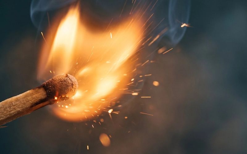 A single wooden match striking into a bright amber flame in the dark -- the spark of an idea AI can't copy.