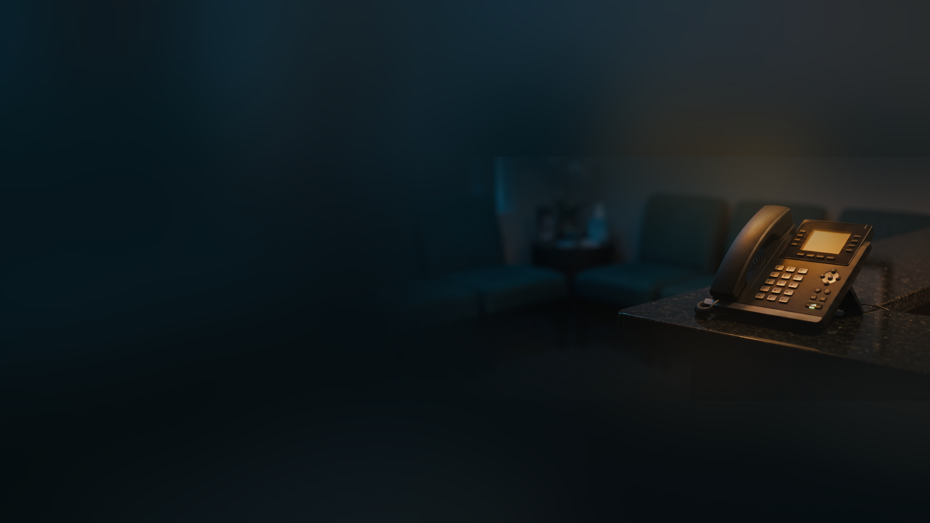 An office desk phone with its screen lit on a reception counter, an empty waiting room blurred behind it