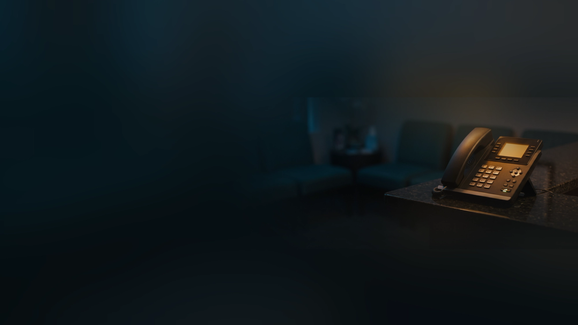 An office desk phone with its screen lit on a reception counter, an empty waiting room blurred behind it