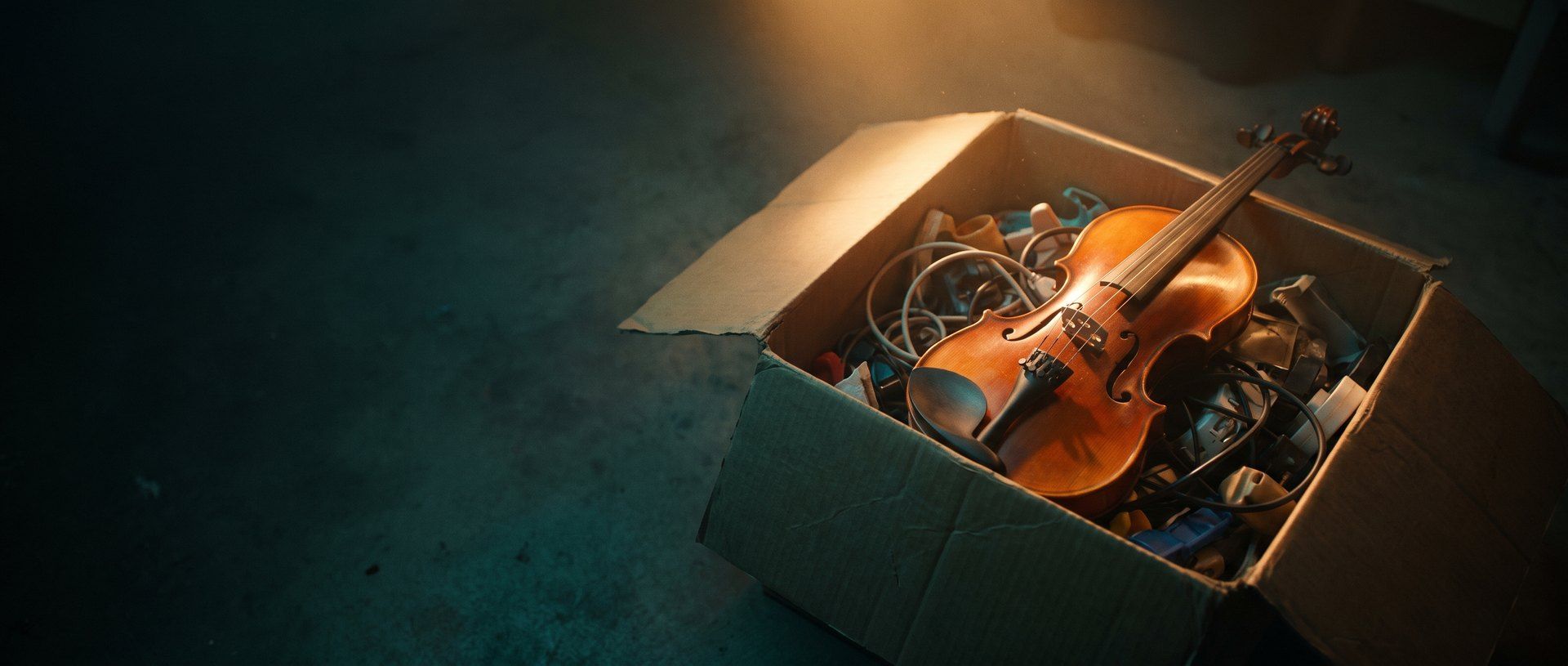 A handcrafted violin with glowing varnished wood lying across an open cardboard box of tangled cables and cheap plastic junk on a dark floor, lit by a single warm amber light.