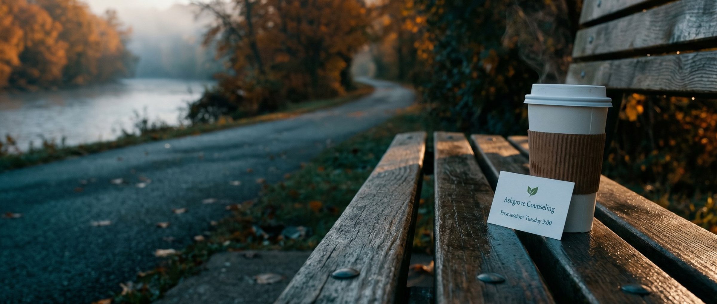 Early morning on a wet wooden park bench beside a misty river trail, a paper coffee cup with a small card tucked against it that reads Ashgrove Counseling, First session: Tuesday 9:00