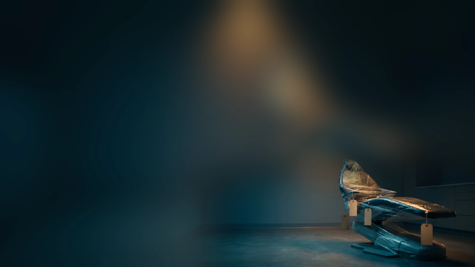 A brand-new dental chair still wrapped in factory plastic with blank shipping tags, in an empty dental operatory under a single warm light