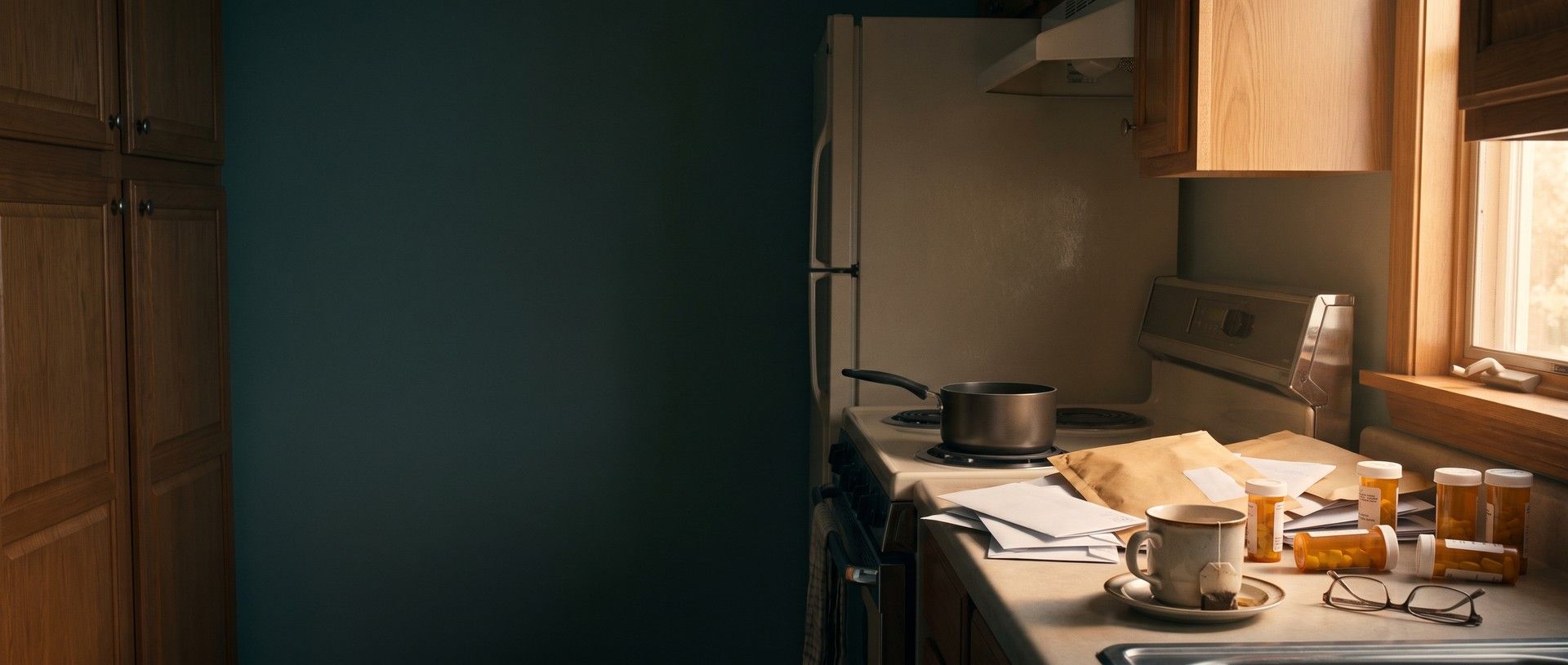 A dated kitchen in morning light: a pot left on the stove, a pile of unopened mail, a cold cup of tea, reading glasses and scattered pill bottles on the counter