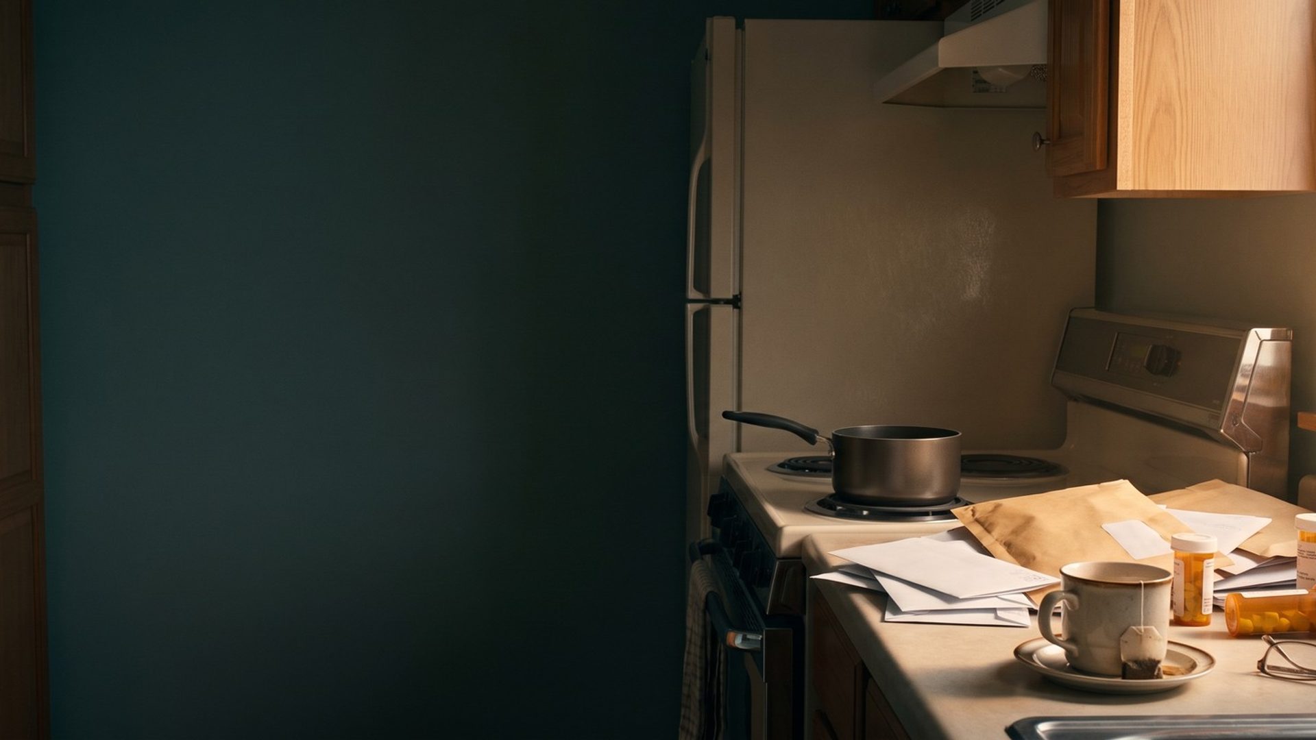 A dated kitchen in morning light: a pot left on the stove, a pile of unopened mail, a cold cup of tea, reading glasses and scattered pill bottles on the counter