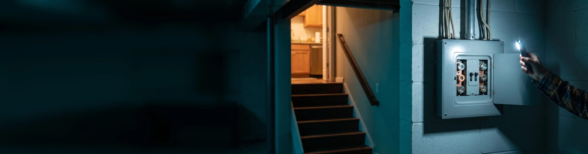 A homeowner's hand holding up a phone flashlight to an old open fuse box with round glass fuses on a basement block wall, warm light spilling down the stairs behind