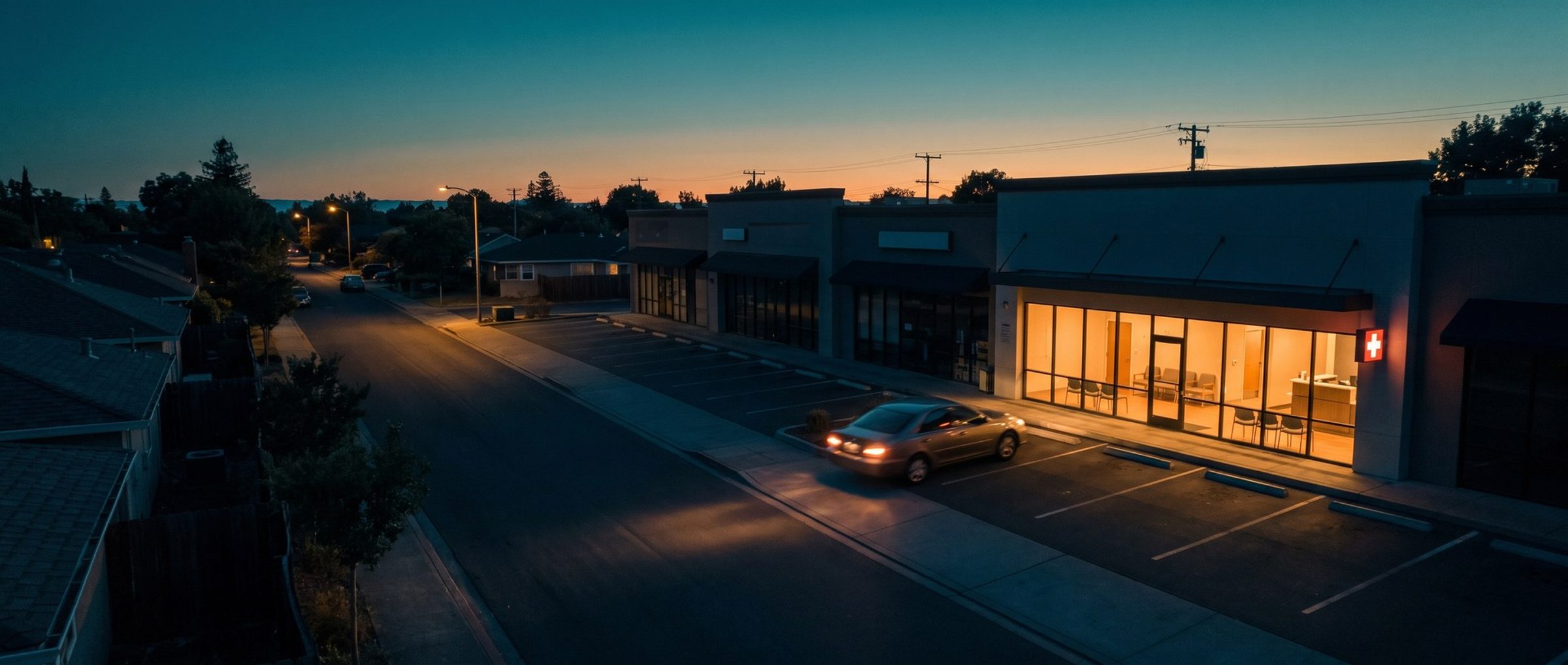 A high view at dusk over a row of small shops, all dark except one urgent care clinic glowing warm behind its glass front with a red cross on the wall, as a single car pulls into its lot