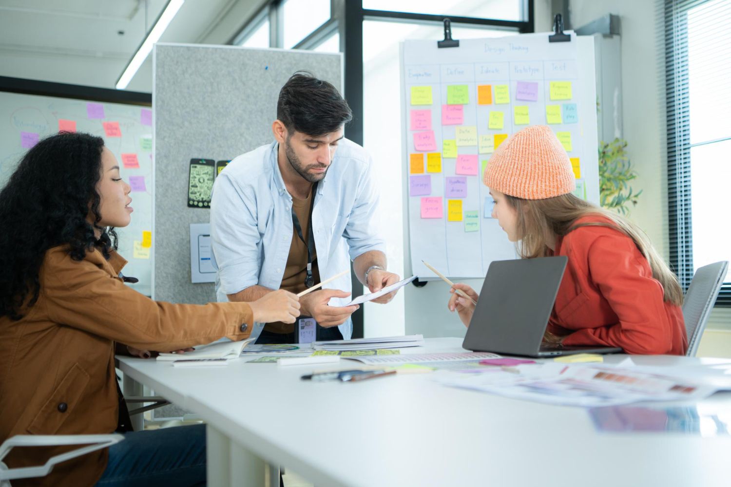 Three colleagues reviewing documents at a table in front of a design-thinking board of sticky notes