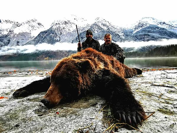Hunter shown with brown bear in front of snow-covered mountains during an Alaska big game hunt.