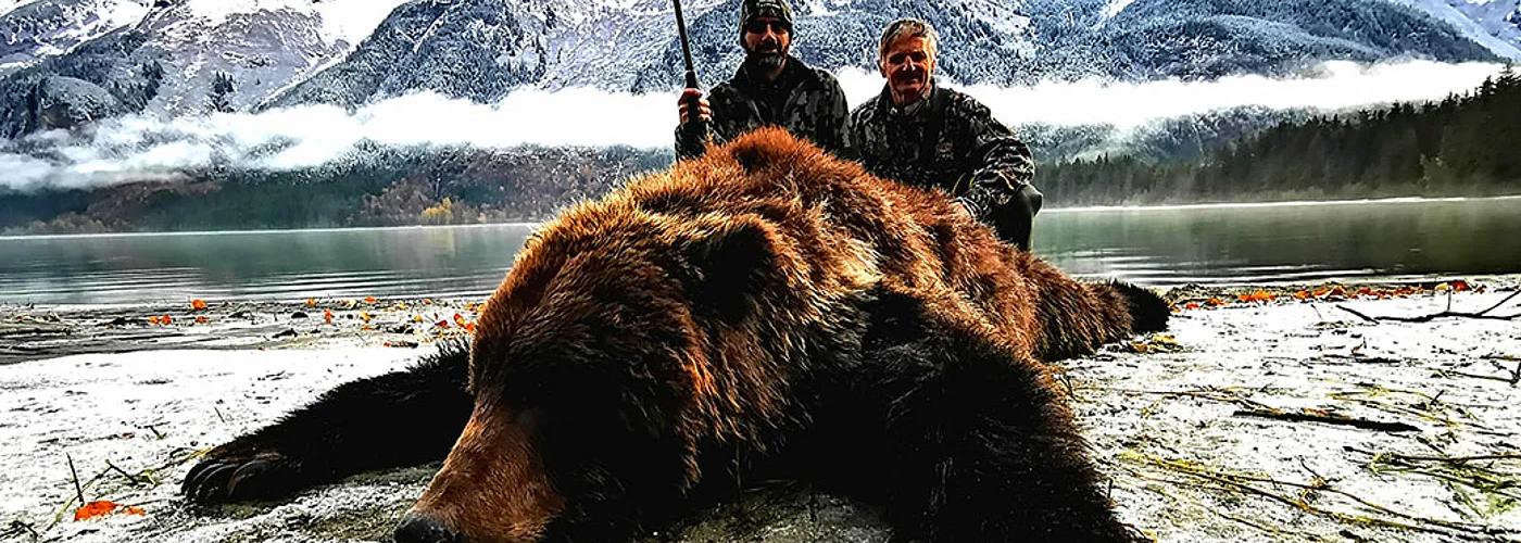 Hunter shown with brown bear in front of snow-covered mountains during an Alaska big game hunt.