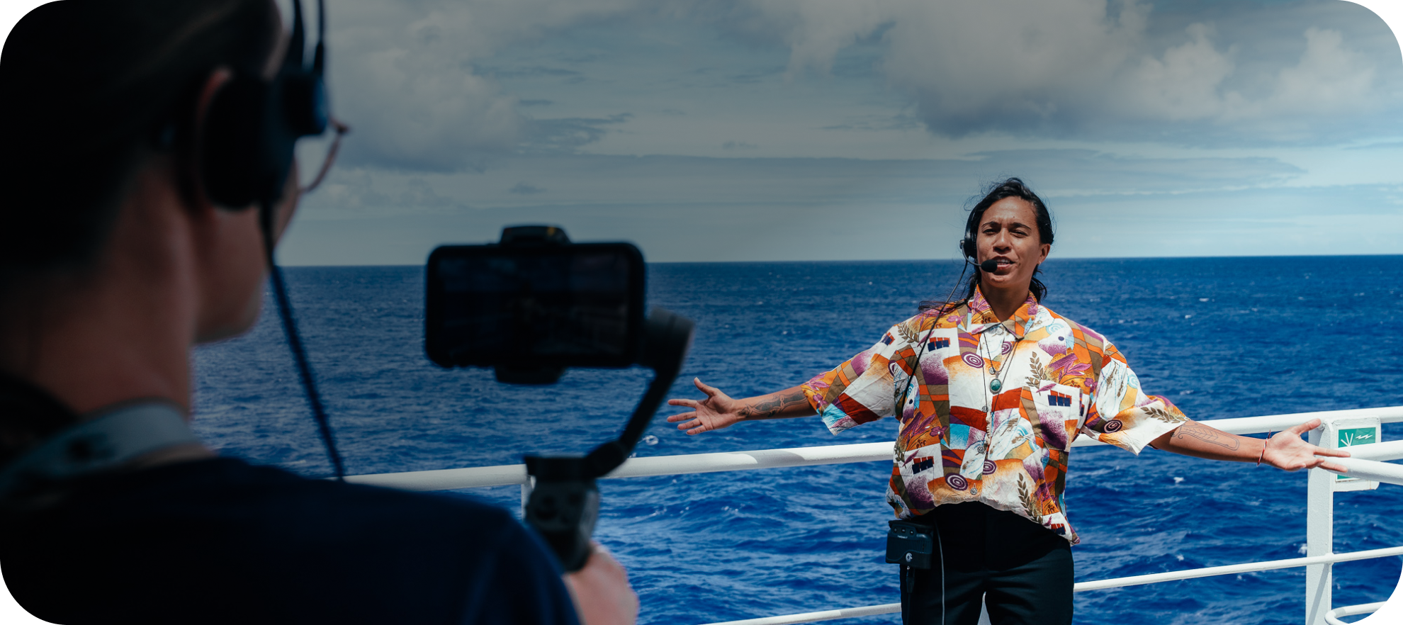 A person wearing a orange graphic button down standing in front of the ocean talking to someone holding a camera. Their arms are out stretched.
