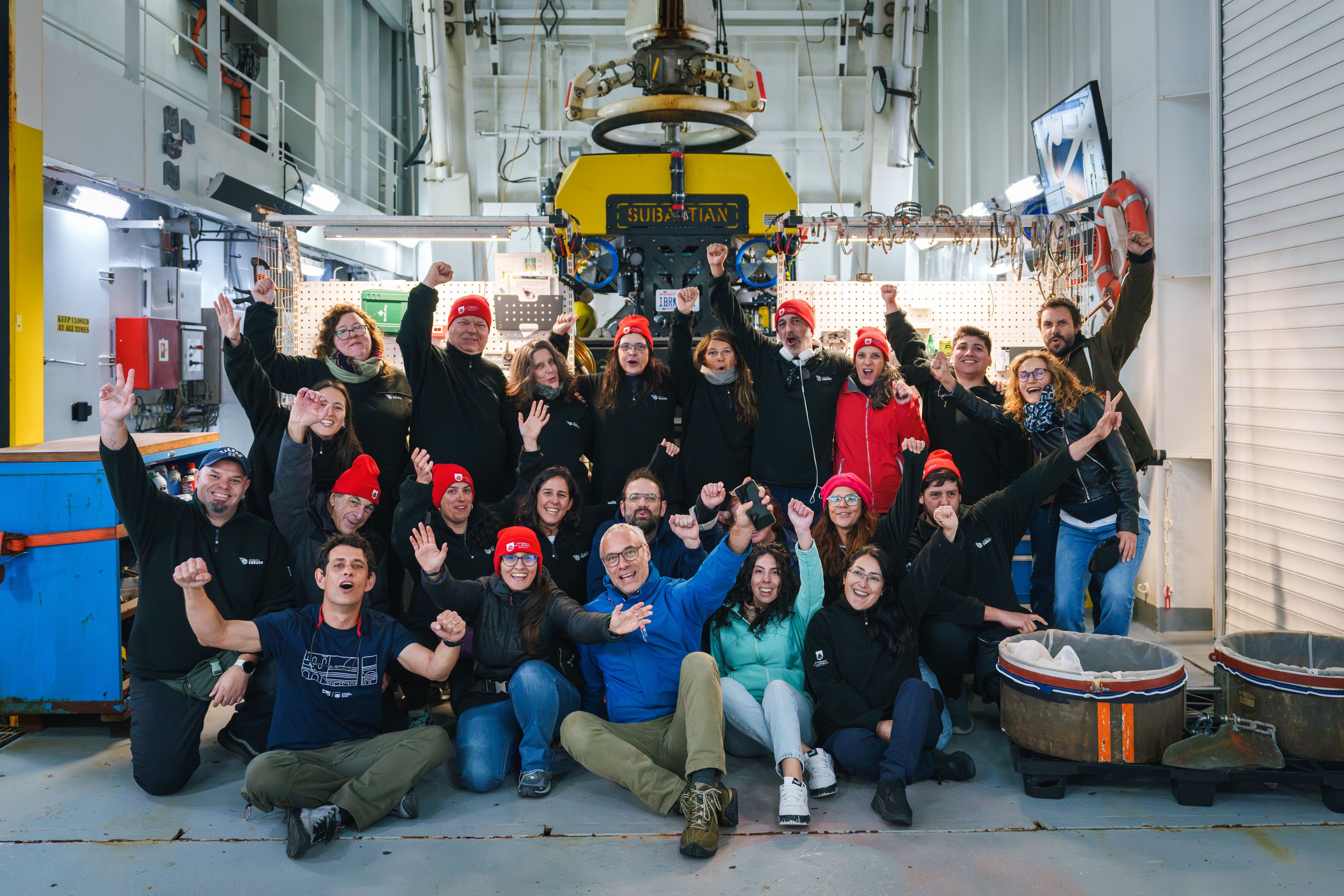 A groupr of researchers raise their hands in celebration in front of a yellow underwater robot. 