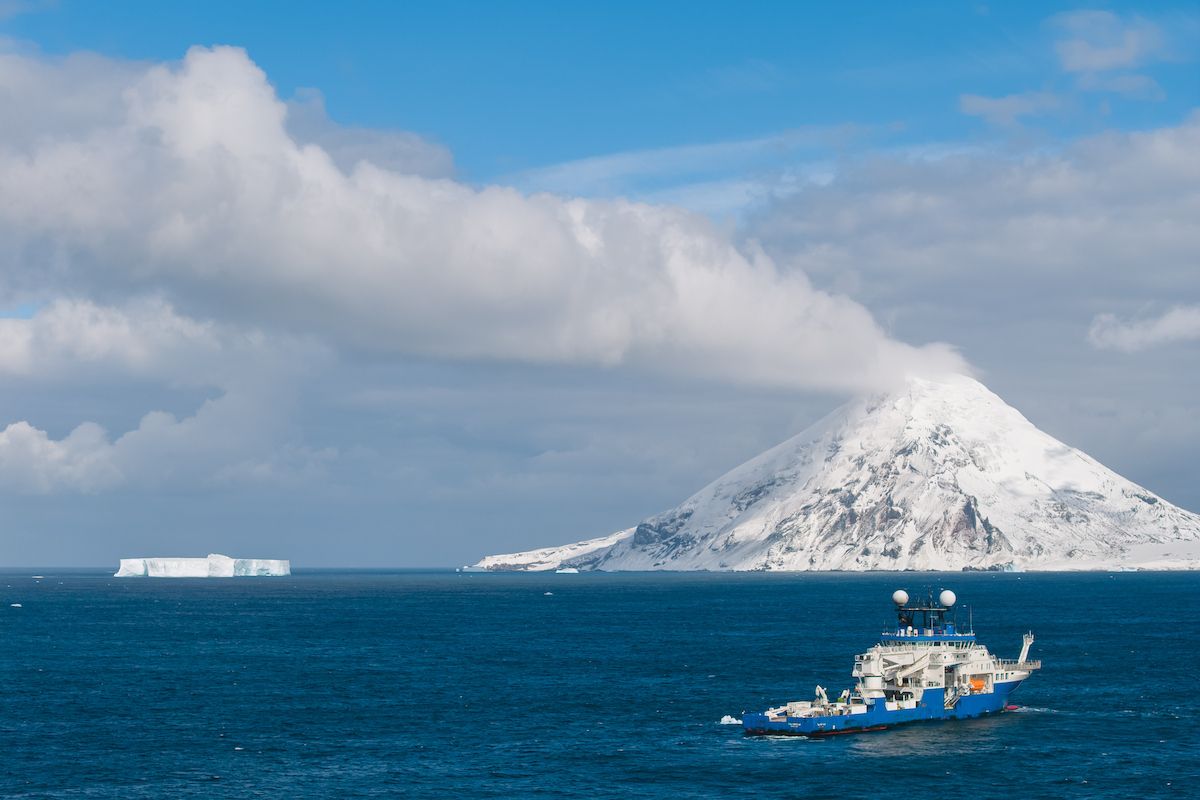 Research vessel Falkor (too) navigating dark blue ocean waters near a snow-covered volcanic peak on Montagu Island under a cloudy sky.
