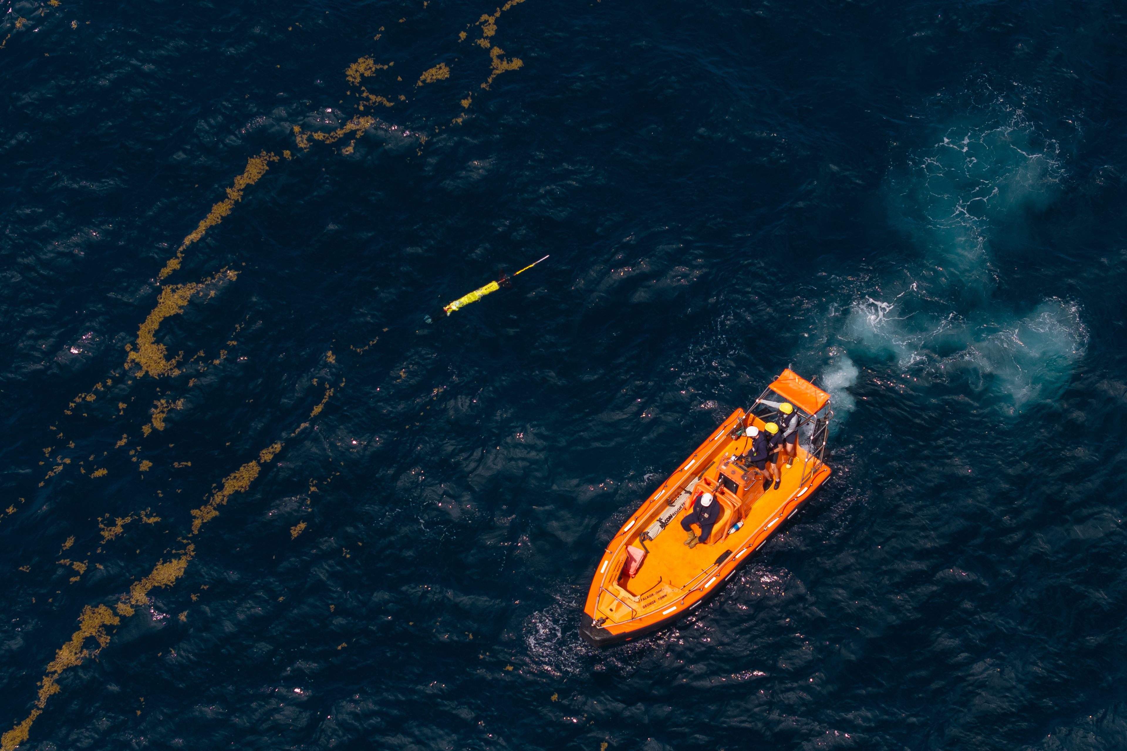 An aerial view of an bright orange boat in deep blue ocean water near a yellow underwater technology and patches of brown sea kelp.