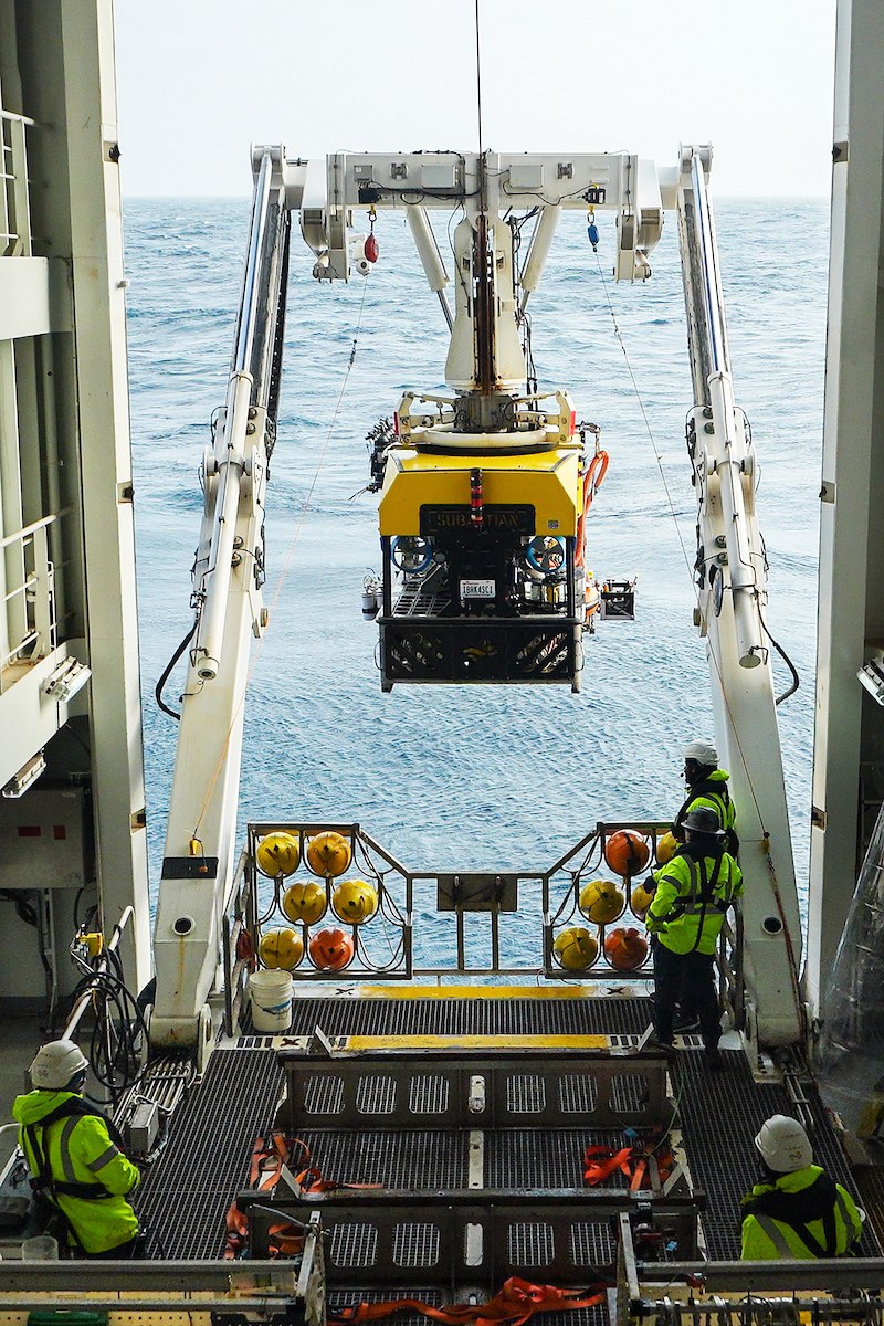 Yellow ROV SuBastian suspended by a large A-frame crane over the open sea as crew members in high-visibility safety jackets observe from the deck.