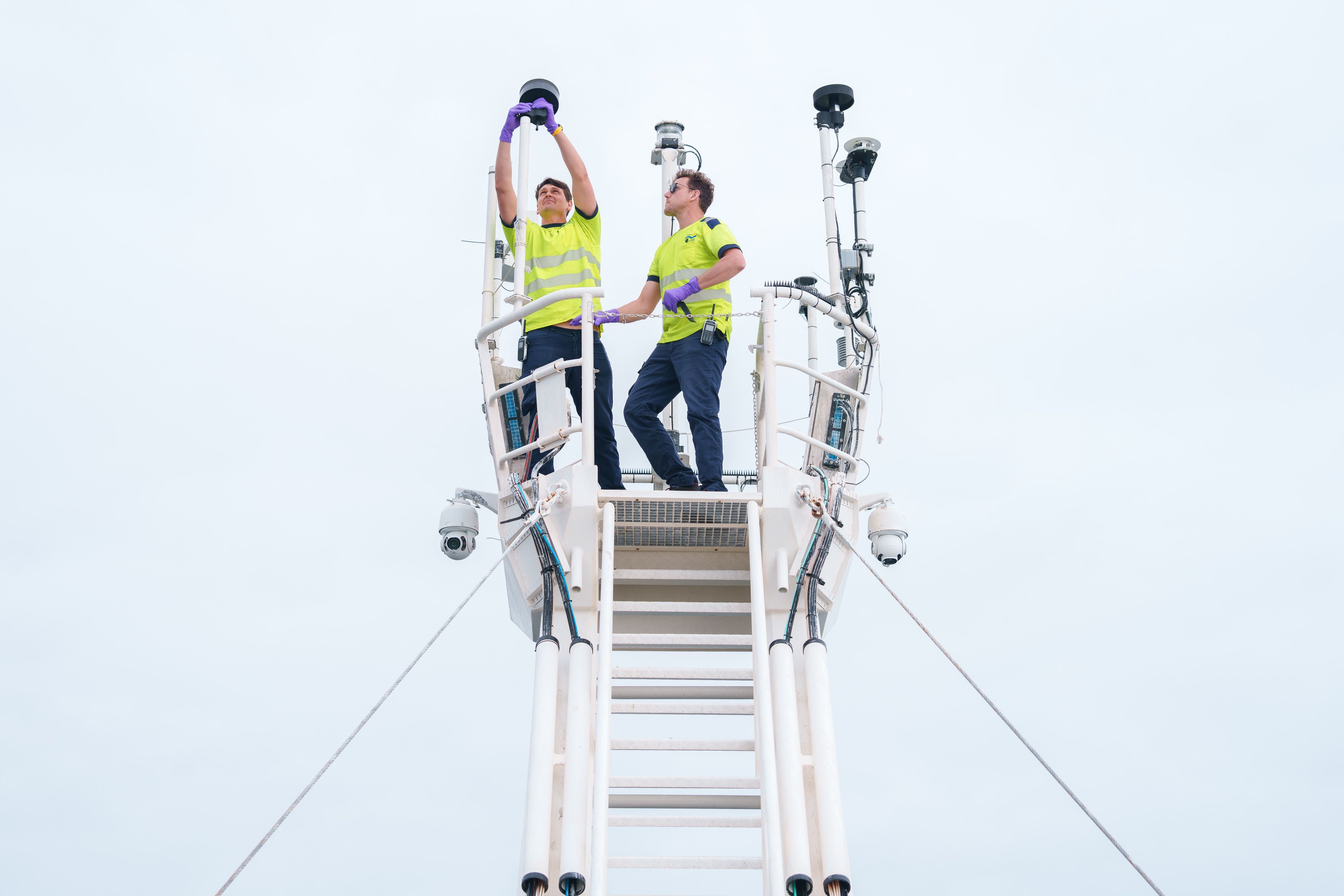 Two technicians wearing yellow on the foremast servicing the met sensors. 