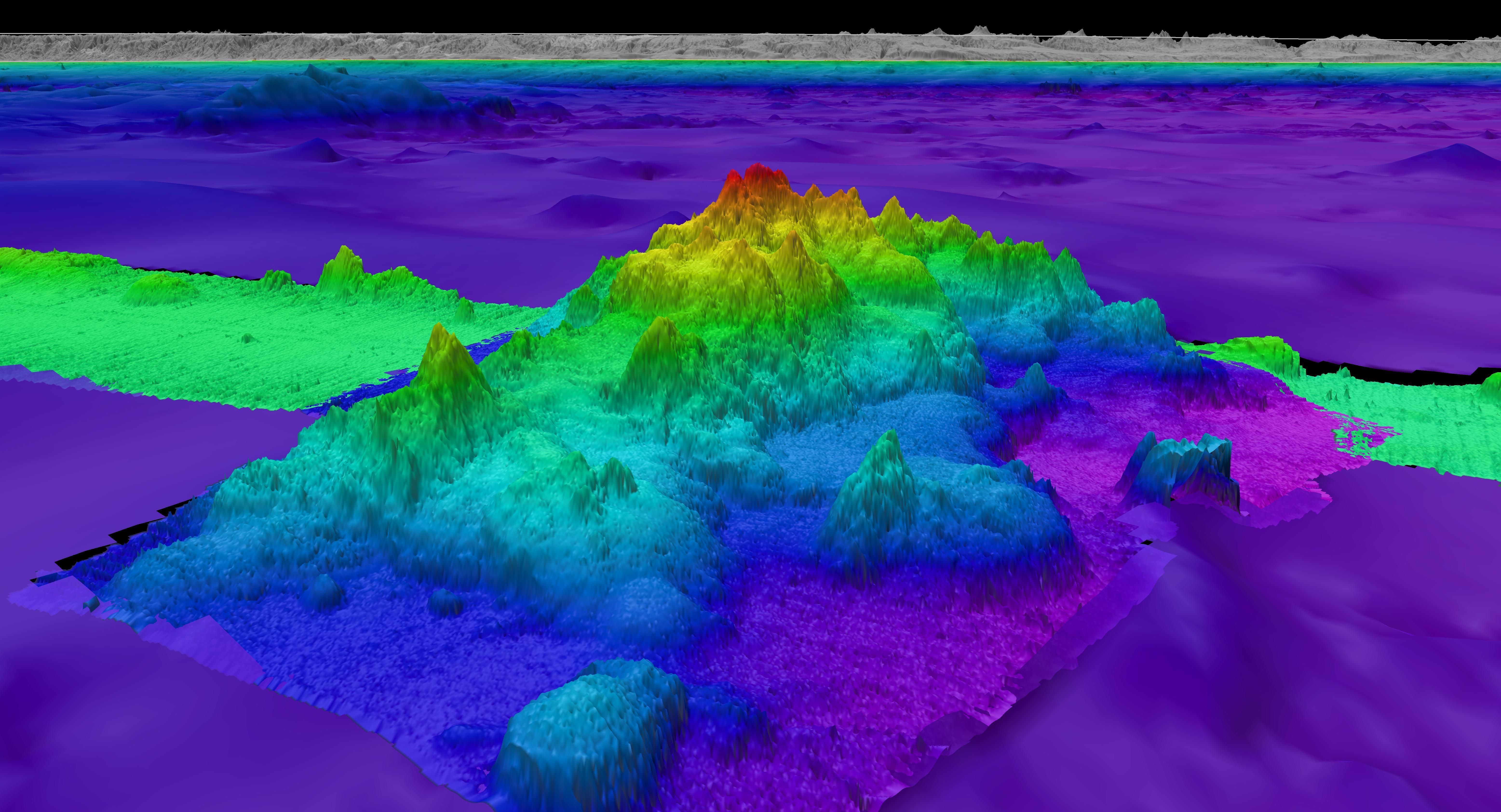 Side view of a subsea seamount with several sharp protruding ridges with a rainbow color scheme mapped by Falkor (too).
